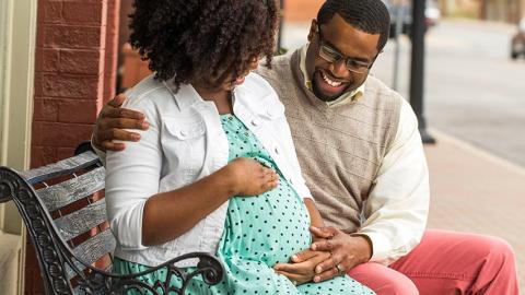 A candid stock photo of a black couple seated on a bench smiling together about the impending birth of their child