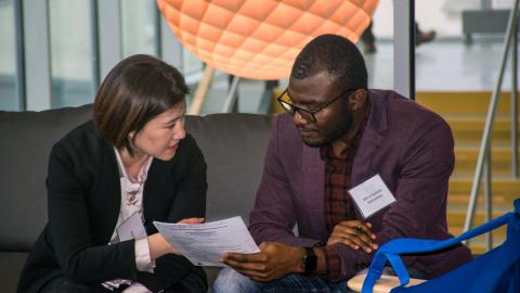 A woman in a dark blue blazer and a man in a purple blazer sit on a couch looking at a document and talking