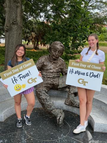 two young women standing on either side of a statue of Albert Einstein. They are holding signs that say First Day of Class Hi Mom & Dad