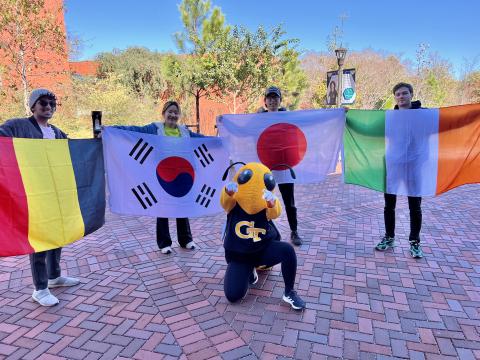 Four Tech students are standing outside on a bright day holding up flags from Spain, Korea, Japan, and Ireland in a semi-circle with Buzz kneeling and pointing at the photographer in the middle, foreground
