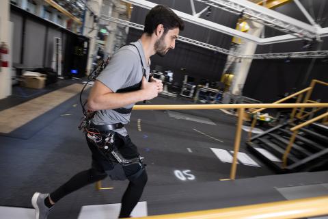 A man wearing a robotic exoskeleton on his upper legs and hips walks up a ramp in a large, open lab space.