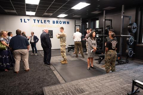 A group of people, some in military uniforms, talk inside an exercise room with dark walls, carpet, and ceiling tiles and a sign on the wall reading "1LT Tyler Brown Fitness Facility."