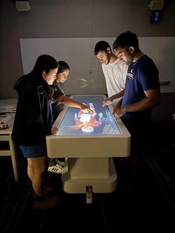 Four students huddle around a lighted table with a virtual human body on it.