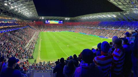A stadium full of fans to watch a nighttime soccer match between the U.S. and Mexico's men's teams. Red and blue lights illuminate the crowd in the stands.