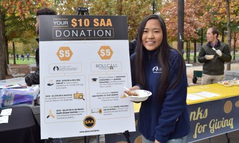 A student stands in front of a sign detailing how a $10 SAA donation is used.