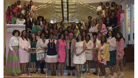 A large group of a large group of Georgia Tech’s Alpha Kappa Alpha Sorority, Incorporated Nu Beta Chapter alumnae stand on two half-circle, glass staircases and wear the AKA colors of pink and green