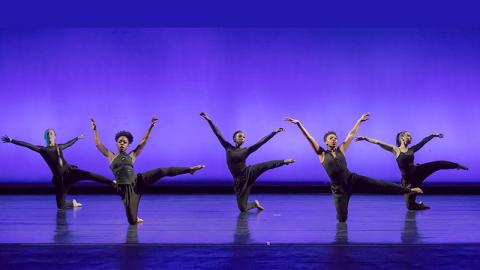 Five female dancers, bare feet, are dressed in black leotards with black pants. They balance on one knee on the ground, while the other leg and arms are outstretched. A vivid purple background is behind them.