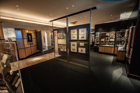 View of museum main gallery. Artifacts are in cases on dark walls. A wooden walkway leads to the center of the room, and the carpet and walls are dark. 