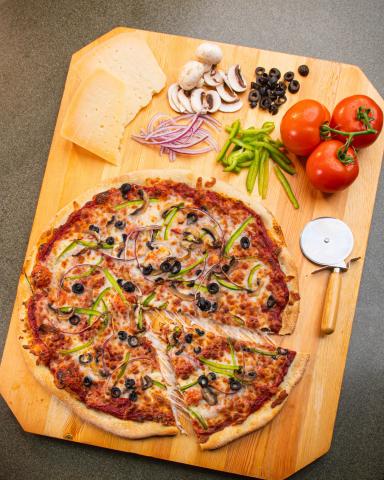 Photo of pizza on a cutting board with cheese, vegetables, and a pizza cutter.