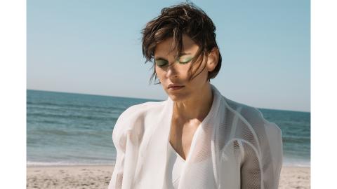 A woman standing on the beach, seen from the chest up, with sand and ocean visible behind her. She is wearing all white, her eyes closed, her hair blowing in the breeze, a bright light reflected on one side of her face.