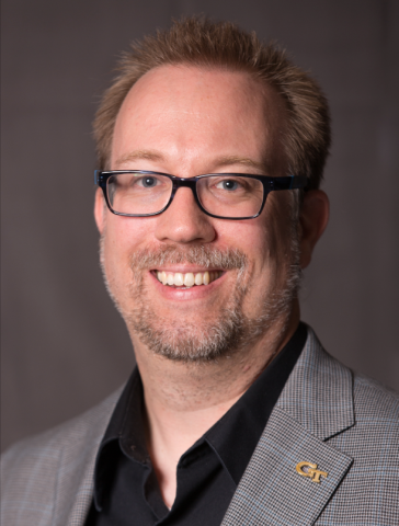 Photo of a man smiling at the camera. He is wearing black glasses and has a goatee. He is wearing a gray blazer over a black shirt. 