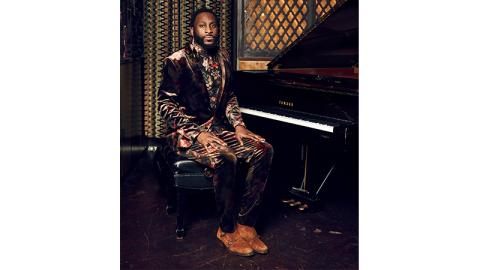 Michael Mwenso wears a brown velvet floral suit. He sits at an open grand piano, hands on his knees, while he looks straight at the camera.