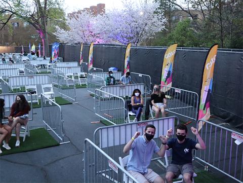 from the viewpoint of standing on a stage the image is of a corner of a parking lot full of audience pods, chairs on a strip of green turf surrounded on three sides by metal bike racks. Behind the smiling students, all wearing face coverings, you see cherry trees up lit in blues and pinks.