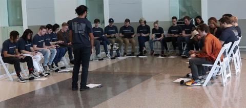 In a large, open room, casually dressed young men and women sit in a semicircle with laptop computers on their laps, intently looking at their screens. A person is standing at the center, conducting.