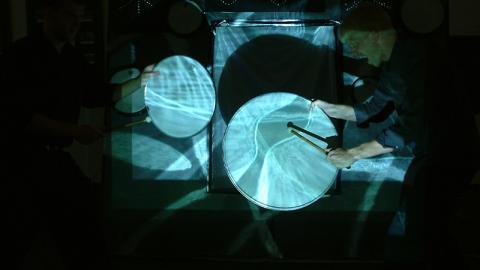 A projection of watery blue patterns of light are cast on two drum heads, each being held vertically and struck with a drumstick.