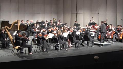 A full orchestra is arrayed on a stage, chairs in four rows stretching from edge to edge of the stage. 
