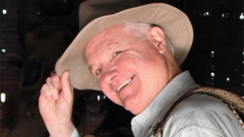 A close up image of a man’s face in three-quarter profile, smiling as he touches the brim of his hat. He seems to be dressed for travel and adventure.