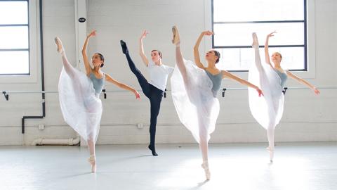 Four dancers are mid-movement, their right legs lifted and toes pointed towards the sky. The three women wear pale blue leotards with gauzy white skirts. The man wears a white T shirt and black tights.