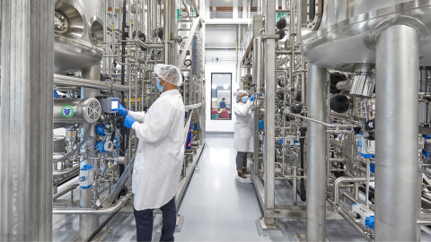 Two people in lab coats and hair covers working in a large lab with lots of metal pipes and containers surrounding them.