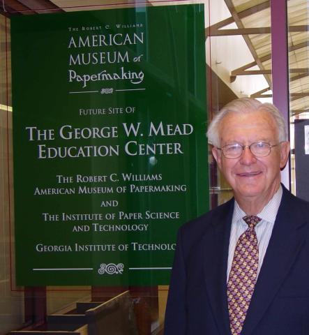 Older man in suit, smiling, stands next to a green sign with white text. 