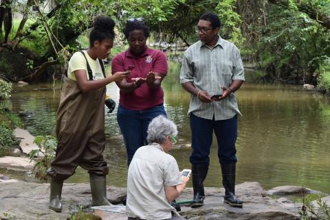 A student stands in waders by a river, consulting with adults and taking pictures of what's in one of the adult's hands.