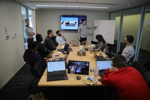 A group of seven students sits around a long conference table with laptops in front of them while another student stands at the front of the group pointing to a white board with words on it. A male instructor is standing on the left side near the door of the conference room.