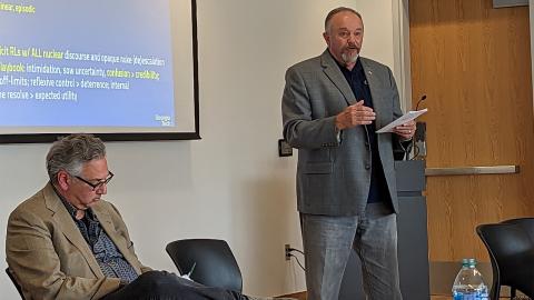 A man dressed in a sportscoat stands and speaks while a colleague takes notes.