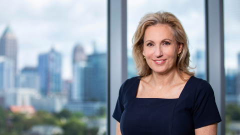 Laura Taylor, a blonde woman with a fair complexion, stands in a navy dress in front of a large window with a blurry skyline of Atlanta visible through the window.