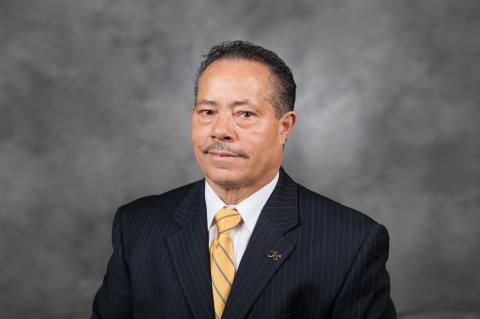 Dr. Archie W. Ervin is sitting in front of a grey and white marbled photo backdrop, wearing a white shirt, yellow striped tie, and a GT pin on his lapel. He has a monotone look on his face and is looking directly at the camera.