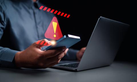 Person holding a cell phone in front of a laptop. Above the cell phone, a large red triangle with a yellow exclamation mark symbolizing a warning sign is digitally displayed.