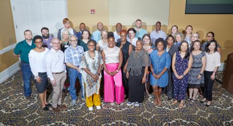 The seventh cohort of the Inclusive Leaders Academy and faculty/staff supporting members stand on patterned carpet, smiling.