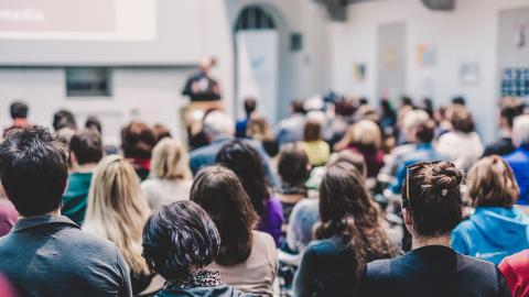 Stock image of people sitting down listening to a speaker at a podium.