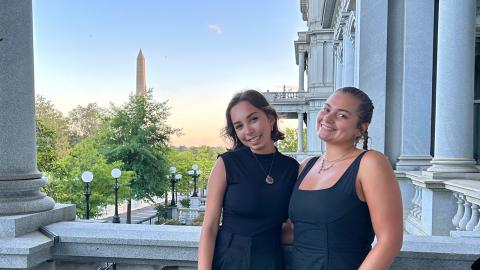 Two students pose for a photo outside an office building with the Washington Monument in the background.