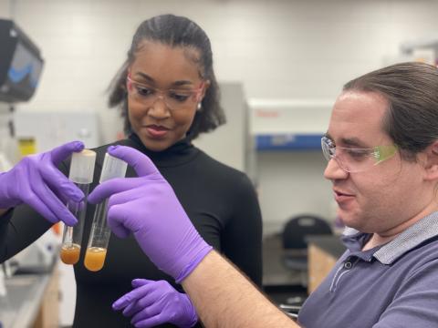 Two people wearing gloves and eye goggles look at substances in a test tube.