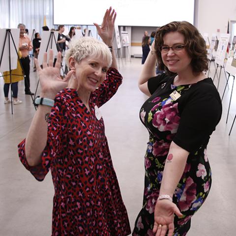 Two women stand facing each other and smiling at the the camera. They stand in front of a poster session and are making a celebratory gesture with their hands and arms.