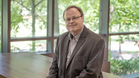 Professor Michael Hoffmann poses for the camera in front of a window wall showing verdant summer foliage.