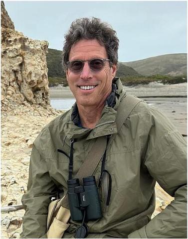 David P. Mindell smiling next to a large rock at the beach with binoculars around his neck.