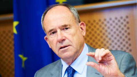 Robert G. Bell gestures during a presentation; the European Union flag is in the background.