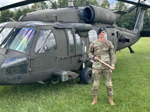 Mason Ladd stands in uniform holding an axe in front of a military helicopter.