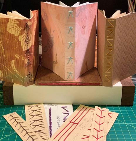 Three neutral colored books with creative bindings, stacked on a larger book that is flat underneath. Bookbinding sewing patterns displayed in front of books.