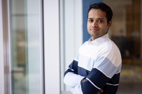 A young man in a collared shirt with blue stripes folding his arms and smiling at the camera