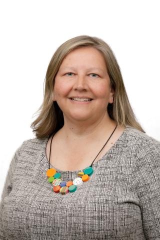 A woman with shoulder-length blond hair wearing a grey top and colorful beaded necklace, against a white background