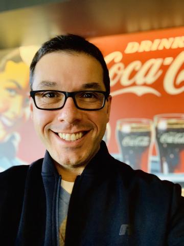 A man with short black hair stands in front of a Coke sign.