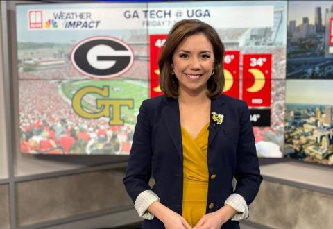 A woman with brown hair sits at a TV anchor desk with weather graphics behind her.