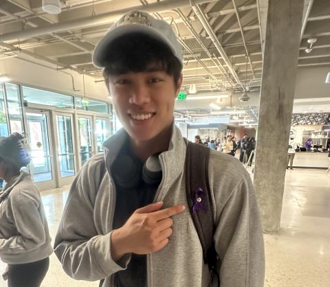 A male student points to a purple ribbon pinned to the strap of his backpack