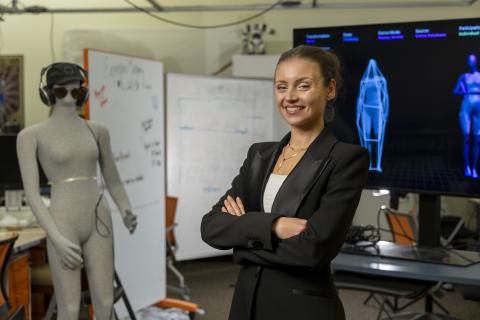female with her hair pulled up and wearing a black suit jacket standing in front of two screen monitors showing blue outlines of human forms