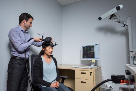 Researcher adjusting a device on another person’s head in a lab, with a computer displaying brain imaging data and a mounted camera in the background.