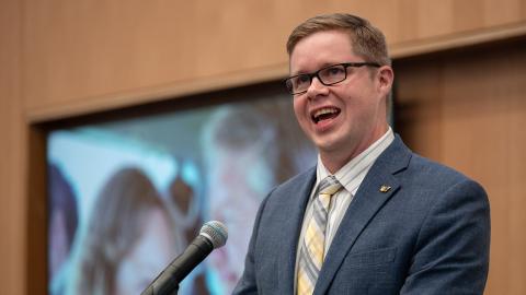 A man in a blue suit and yellow tie speaks at a podium