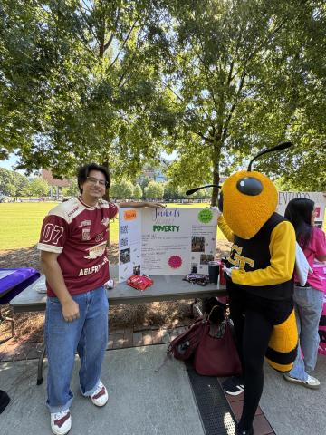 A young man and Buzz stand in front of a Yellow Jackets Against Poverty poster.