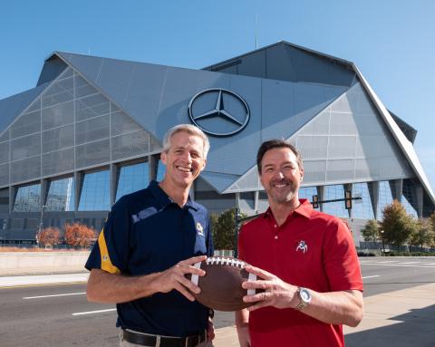 A tall white man wearing a blue GT-branded polo standing next to a slightly shorter man wearing a UGA-branded red polo. They're smiling and both holding a football.